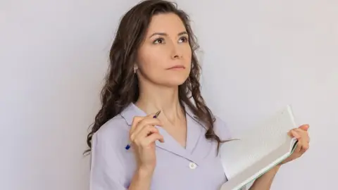 Premýšľajúca žena s poznámkovým blokom a perom, pôsobiaca pokojne a sústredene – symbol organizovanosti a plánovania.