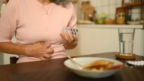 Žena sediaca pri stole si drží brucho a v ruke drží balenie tabliet, pred ňou stojí pohár vody a tanier s jedlom.