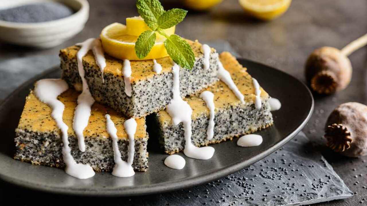 Makový koláč nakrájaný na štvorce s citrónom a jemnou polevou na vrchu, podávaný na tmavom tanieri.