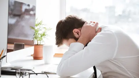 Unavený muž sedí pri pracovnom stole a drží si hlavu v rukách, pôsobí vyčerpaným dojmom.