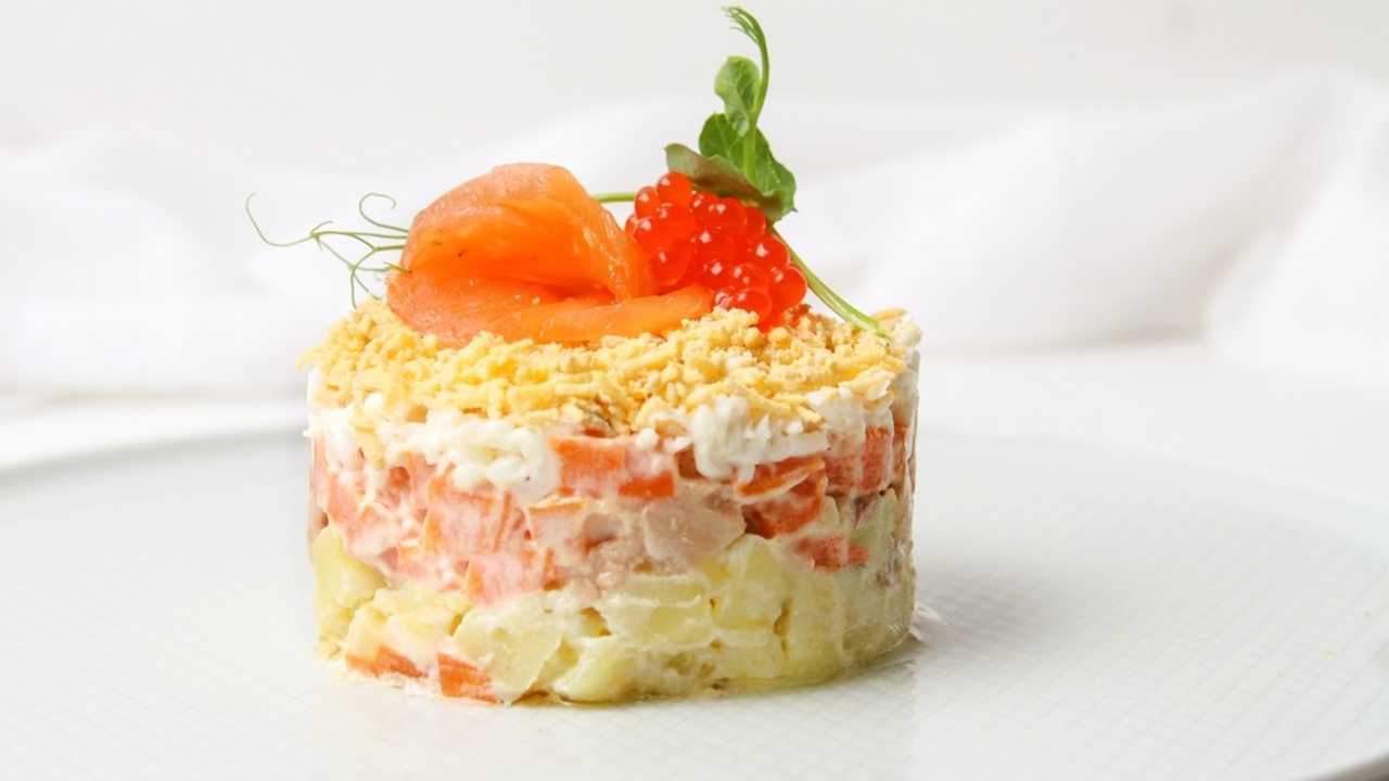 Tradičný ruský vrstvený šalát zdobený plátkom lososa a červeným kaviárom na sivom tanieri s vidličkou a nožom na bielom stole. Bočný pohľad, detail.