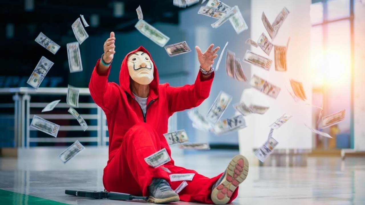 Osoba v červenom kostýme a maske pripomínajúcej seriál Dom z papieru, obklopená lietajúcimi bankovkami.