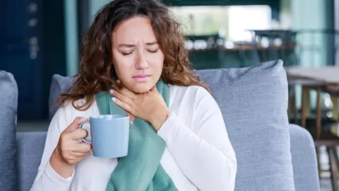 Žena s bolesťou hrdla sedí na gauči a drží v ruke hrnček teplého nápoja.