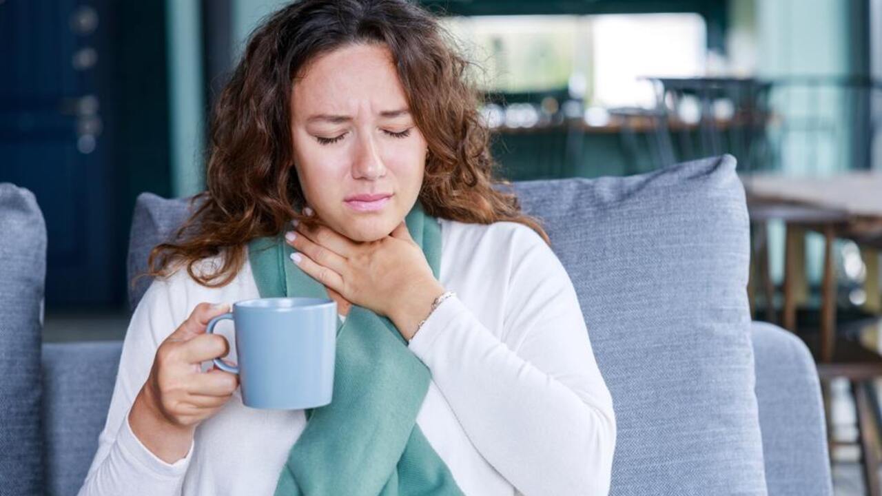 Žena s bolesťou hrdla sedí na gauči a drží v ruke hrnček teplého nápoja.