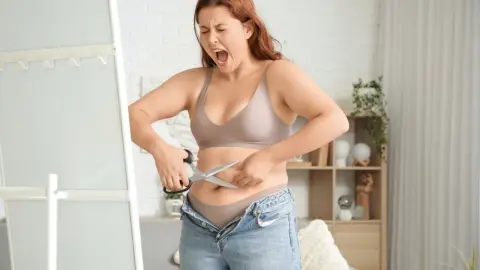Plus size žena v spodnom prádle sa snaží pomocou nožníc prerezať príliš tesné džínsy, ktoré nedokáže zapnúť.