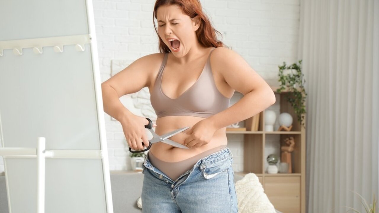 Plus size žena v spodnom prádle sa snaží pomocou nožníc prerezať príliš tesné džínsy, ktoré nedokáže zapnúť.