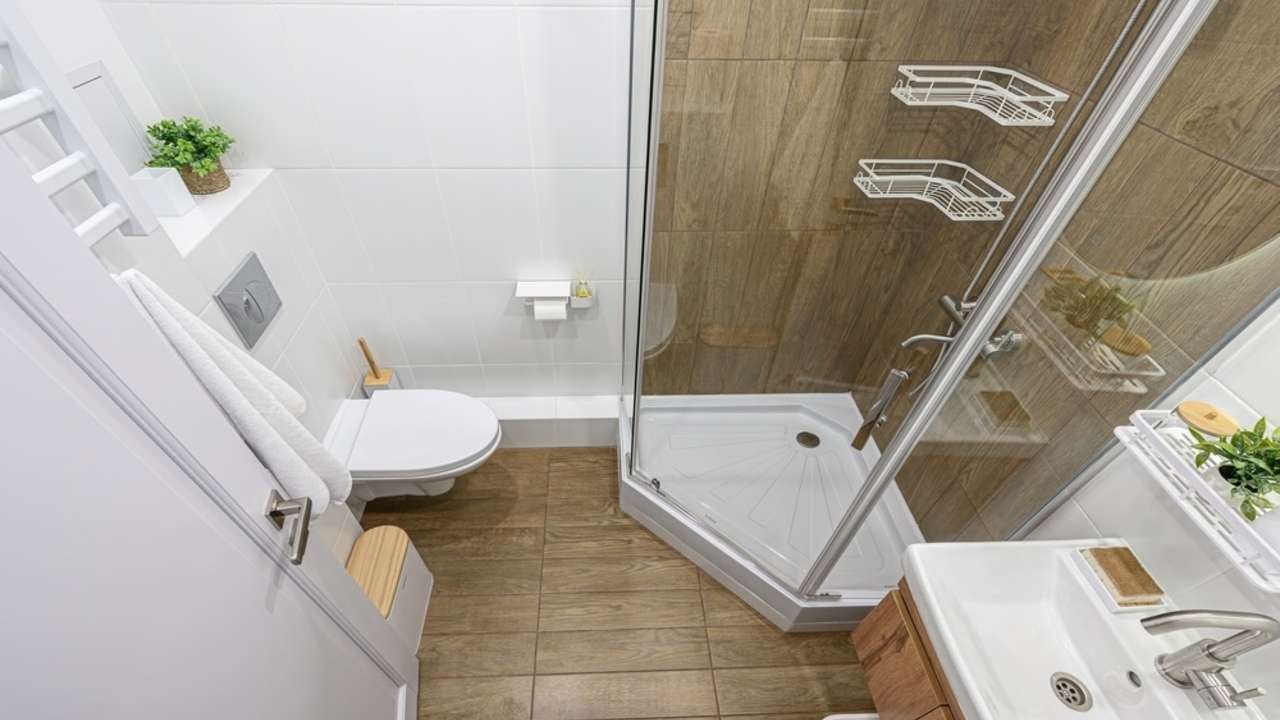 Svetlá malá kúpeľňa s preskleným sprchovým kútom, drevenou podlahou a minimalistickým zariadením, ktoré opticky zväčšujú priestor.