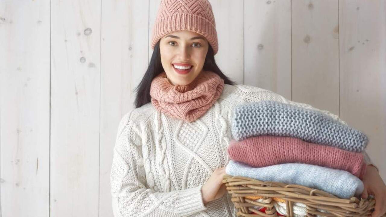 Usmievavá žena v zimnej čiapke a šále drží košík plný zložených pletených svetrov