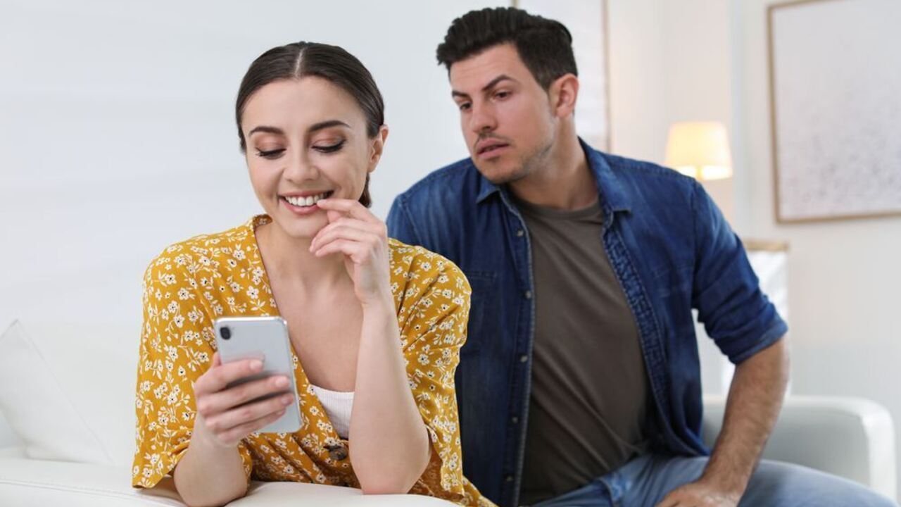 Žena používa mobilný telefón, zatiaľ čo partner za ňou podozrievavo sleduje obrazovku