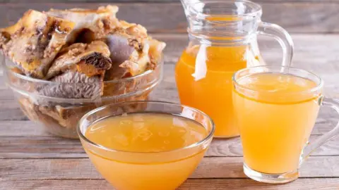 Domáci kolagénový vývar z kostí v miske a pohári vedľa surových kostí na drevenom stole