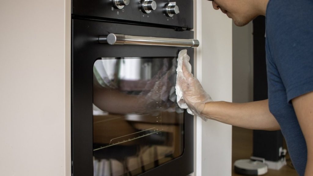 Čistenie sklenených dvierok modernej vstavanej rúry v domácej kuchyni, pričom si má nasadiť ochranné rukavice a zotrieť penu z čistiaceho prostriedku, aby boli dvierka bez jediného fľaku.