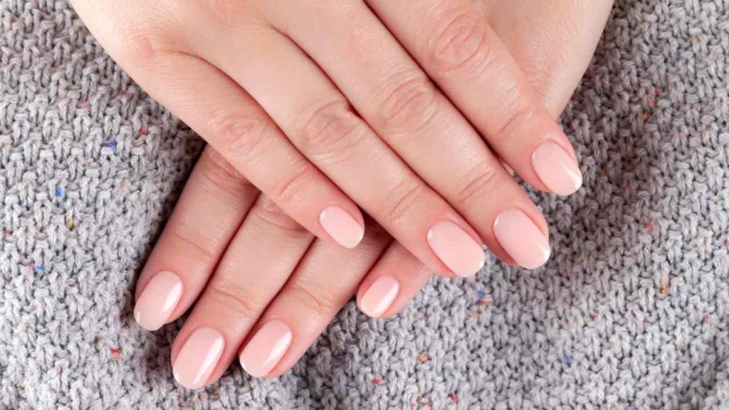 Ženské ruky s krásnou prírodnou manikúrou – ružové nechty v odtieni nude na pozadí zo šedej pleteniny. Koncepcia starostlivosti o nechty