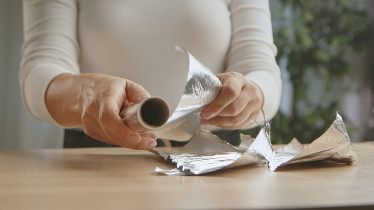 Záber zblízka na ruky ženy, ktorá manipuluje s lesklou rolkou hliníkovej fólie na drevenom stole, čím sa zdôrazňuje činnosť v domácnosti.