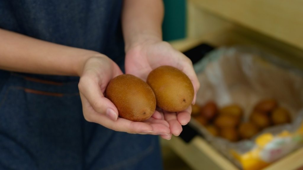 Záber zblízka na ruky ženy, ktorá drží dve zrelé kiwi pred regálom plným ďalších tropických plodov v ovocnom obchode; v prirodzenom svetle je vidieť štruktúru a čerstvosť hnedého chmýrovitého povrchu.