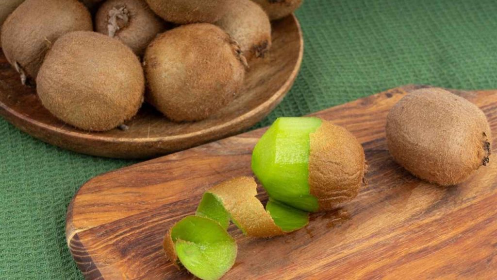 Šupka z kiwi na drevenej doske na krájanie, lúpanie kiwi. Rozrezané kiwi, čerstvé šťavnaté tropické ovocie so semienkami, kúsok kiwi na bielom pozadí