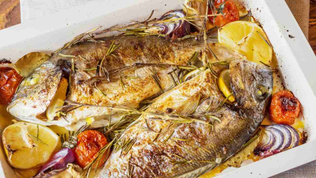 Dve lahodné celé pečené ryby. Pečený dorado s citrónom, cibuľou, rozmarínom, cherry paradajkami a korením na drevenom podklade. Lahodný pečený dorado na plechu. Diétne a zdravé jedlo. Pražma zlatá