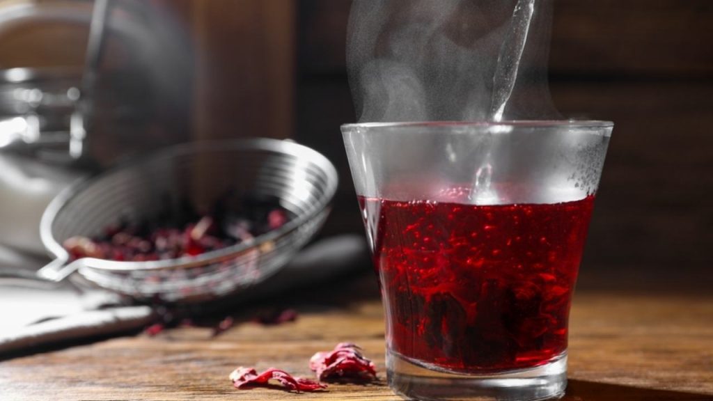 Príprava čaju z ibišteka. Naliatie horúcej vody do šálky s okvetnými lístkami ibišteka na drevenom stole, detailný záber.