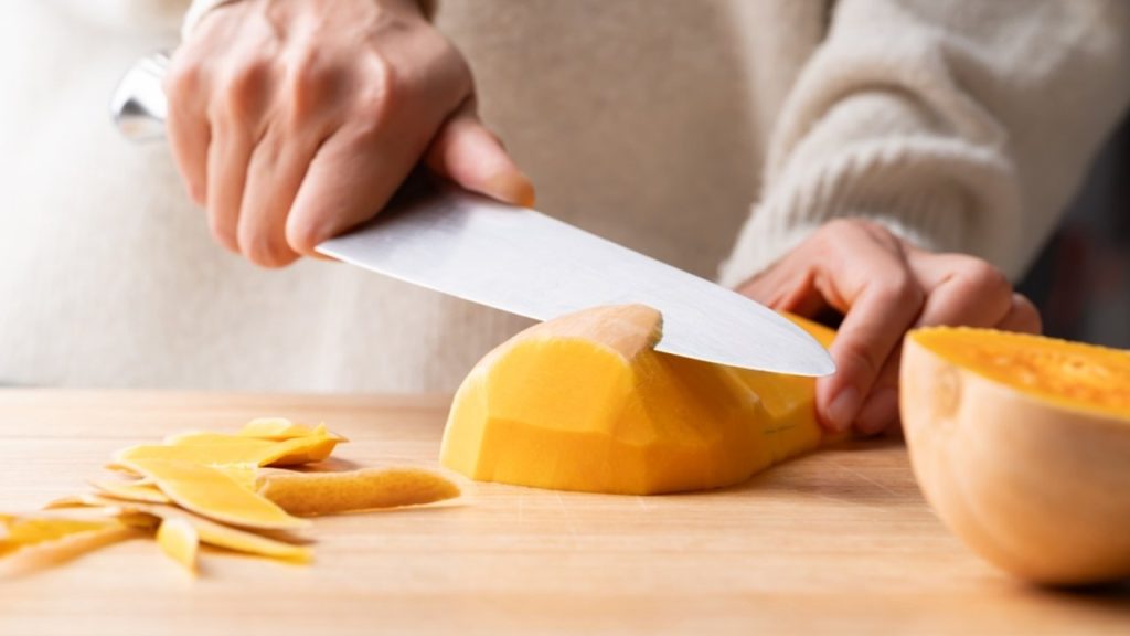 Žena držiaca v ruke kuchynský nôž a šúpajúca tekvicu butternut, Domáce varenie