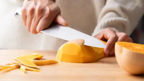 Žena držiaca v ruke kuchynský nôž a šúpajúca tekvicu butternut, Domáce varenie