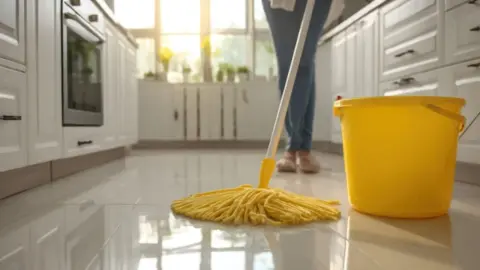 Žena umýva podlahu v modernej kuchyni pomocou mopu a vedra. Podlaha je veľmi lesklá a odráža slnečné lúče, ktoré sem prenikajú.