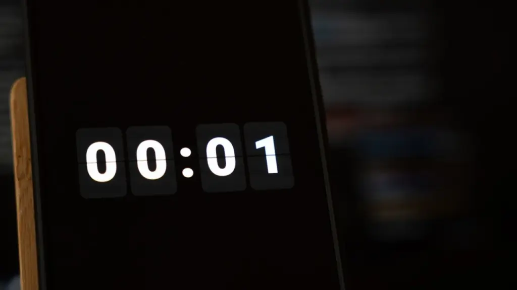 Odpočítavanie poslednej sekundy, ktoré na tmavej obrazovke smartfónu vedľa dreveného stojana zobrazuje biele číslice na otočných hodinách s hodnotou 00:01; symbolika jednej zostávajúcej sekundy, ponuka na poslednú chvíľu, uplynutie termínu a koniec času